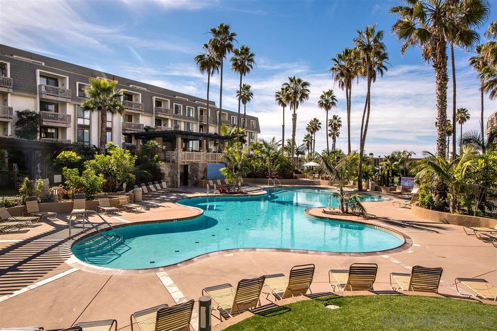 A large curvy pool is surrounded by palm trees and tropical landscaping. Lounge chairs are neatly placed along the pool's edge, inviting relaxation. In the background, a three-story building provides a serene view, enhancing the private atmosphere of the complex.
