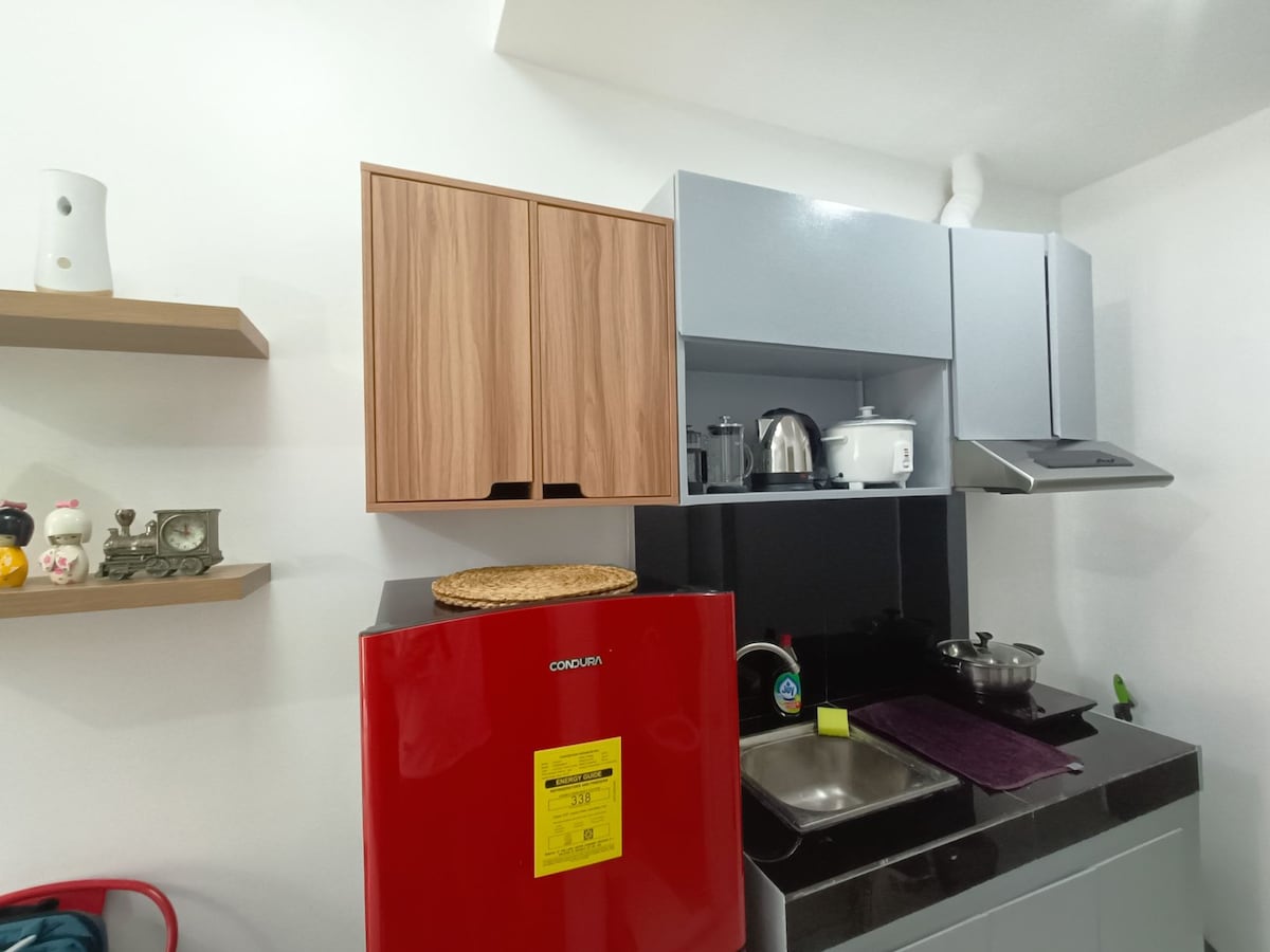 A modern kitchen area is equipped with a red refrigerator and sleek cabinetry in neutral tones. Essential appliances such as a kettle, rice cooker, and induction stove are visible on the countertop, which is complemented by a stainless steel sink and a decorative woven mat.