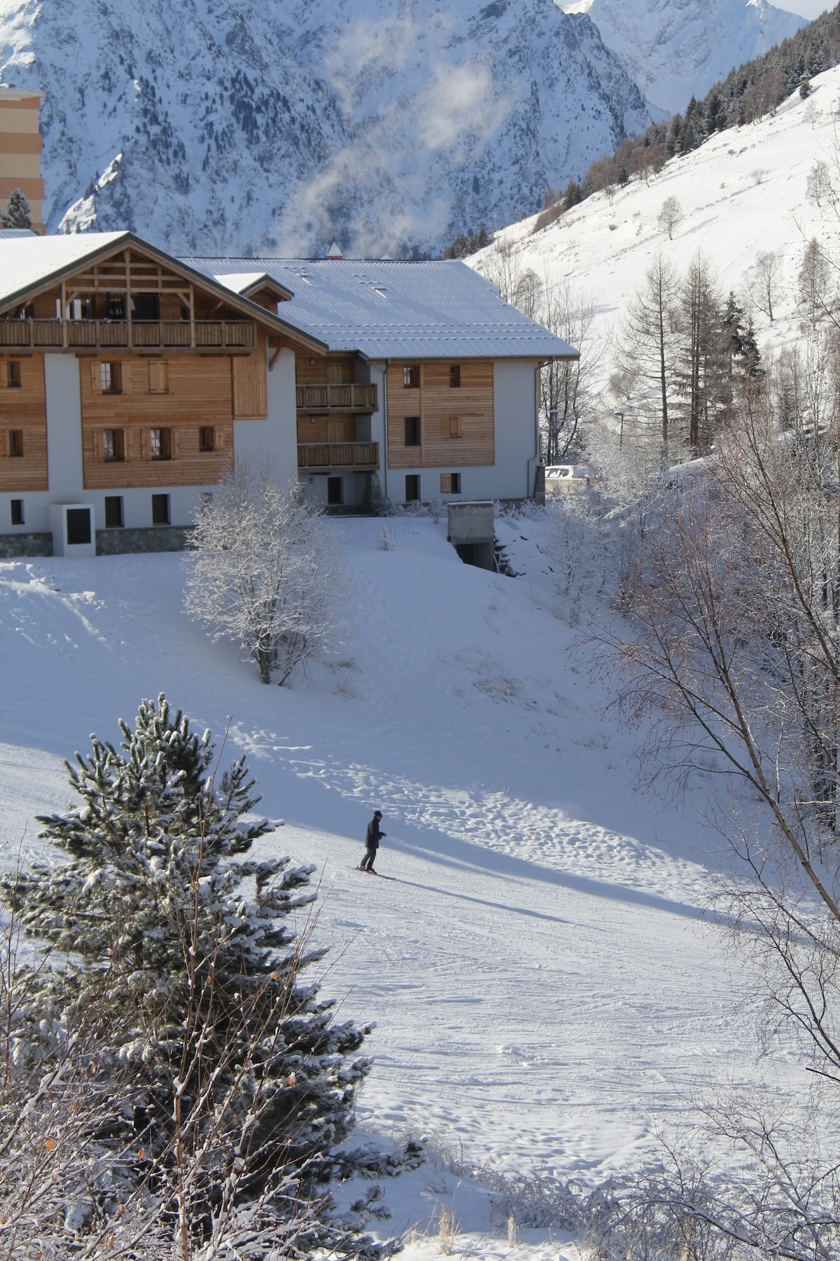 A snow-covered landscape features a modern building with a wooden facade nestled against a mountainous backdrop. A figure skis gently down a slope, flanked by trees dusted with snow, showcasing the serenity of the winter setting.