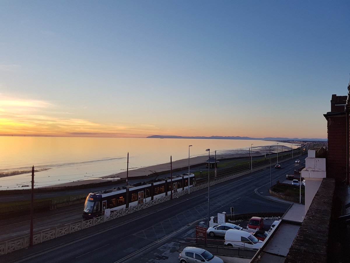 A serene view is presented, featuring a wide expanse of water reflecting the soft hues of sunset. A tram can be seen traveling along the side of the road, with a coastal line visible in the background against a distant horizon.