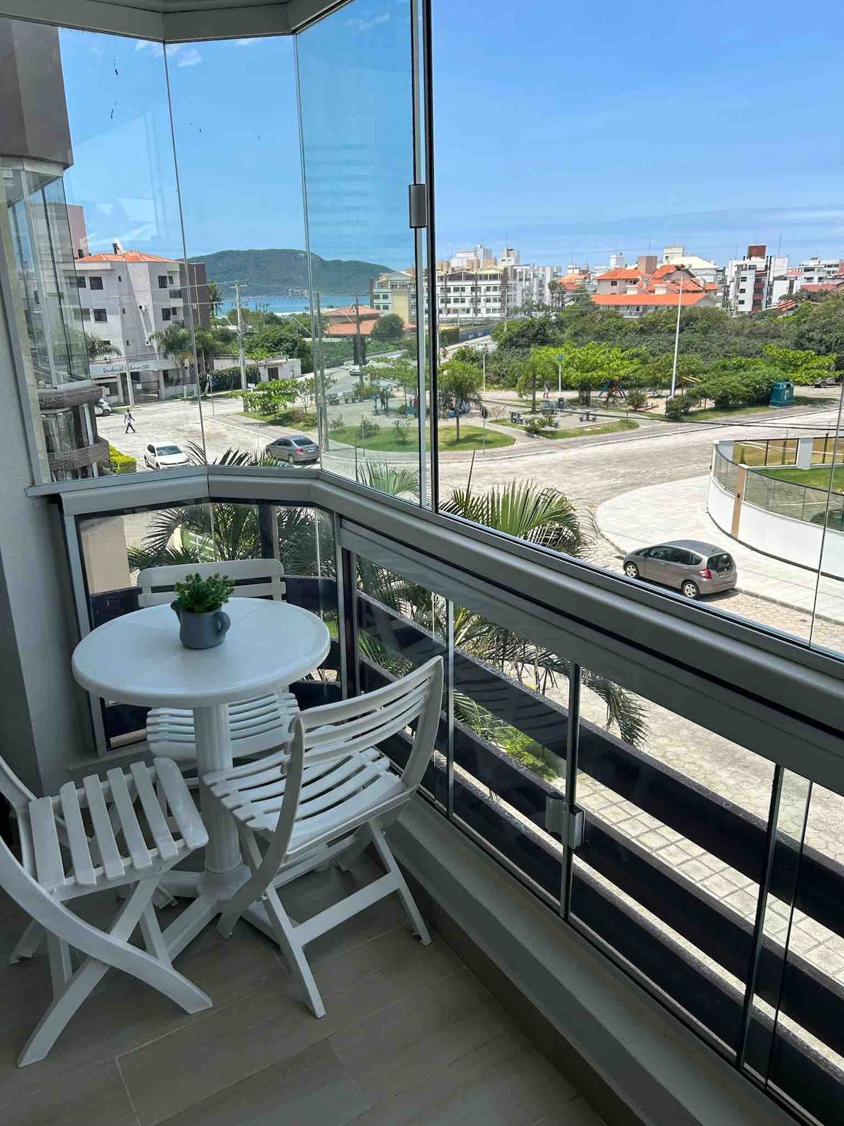 A balcony seating area features a small round table accompanied by two white chairs. The glass railing provides an unobstructed view of the coastline and surrounding trees, enhancing the space with natural light.
