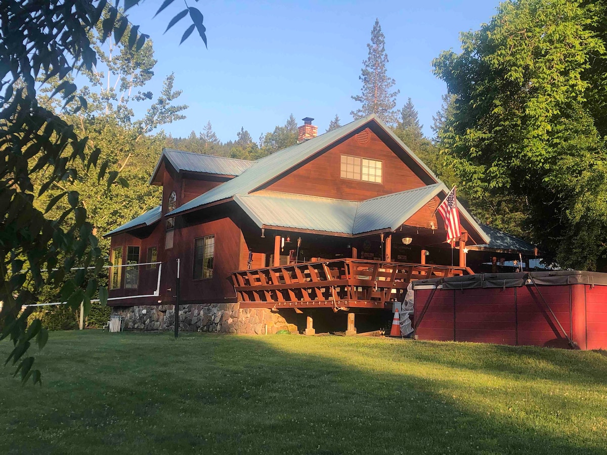 The cabin features a rustic exterior with a wooden facade and a blue metal roof. A large covered porch extends from the front, surrounded by a grassy area. An American flag is displayed, and a swim spa is positioned nearby, blending with the natural surroundings.