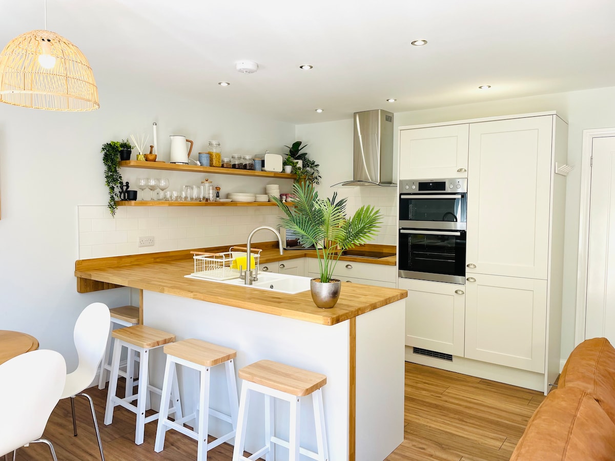 A modern kitchen is highlighted by a spacious island with a built-in sink and wooden countertops. Light cabinetry surrounds the area, complemented by open shelving displaying dinnerware and plants. Bar stools are positioned along one side of the island, with a dining table nearby.