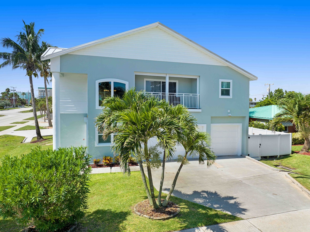 A two-story townhouse is depicted with a light blue exterior and a white balcony overlooking the front yard. Lush green palm trees frame the entrance, with a paved driveway leading up to a single-car garage. Nearby, neat landscaping complements the serene environment.