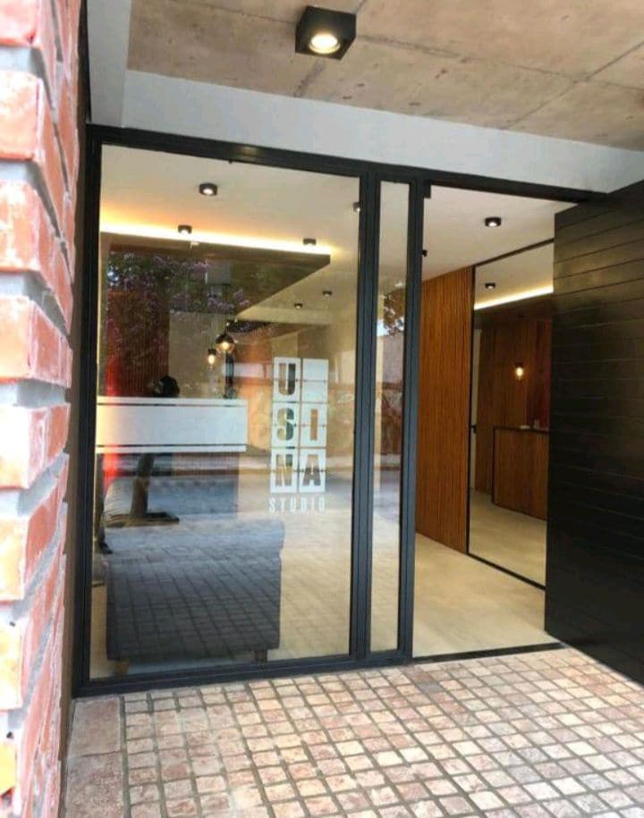 The entrance features a modern glass door framed by a brick wall, displaying the name 'USINA STUDIO.' Inside, warm wood accents and recessed lighting create an inviting atmosphere. The flooring is smooth, and a small reception area is visible through the glass.