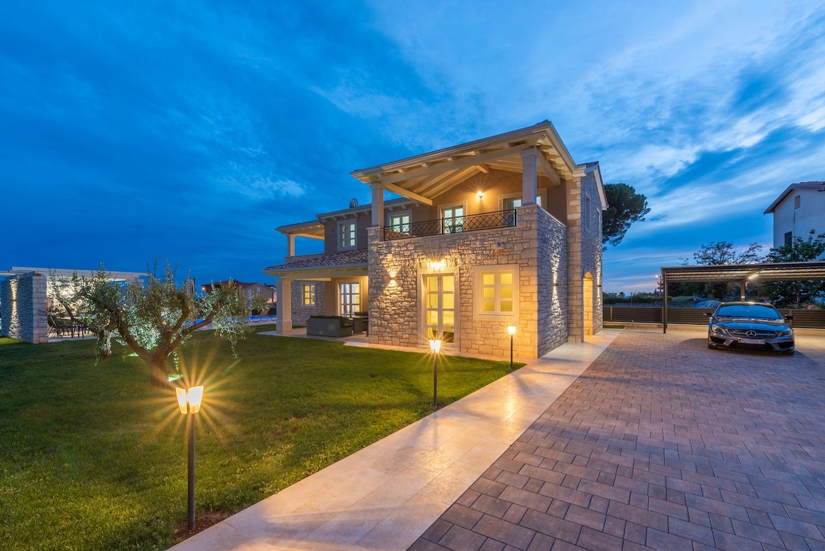 The villa's exterior is showcased during twilight, featuring a modern stone facade and illuminated pathways. A spacious lawn surrounds the building, while outdoor seating areas offer views of the surroundings. A sleek car is parked under a carport, blending functionality with the scenic setting.