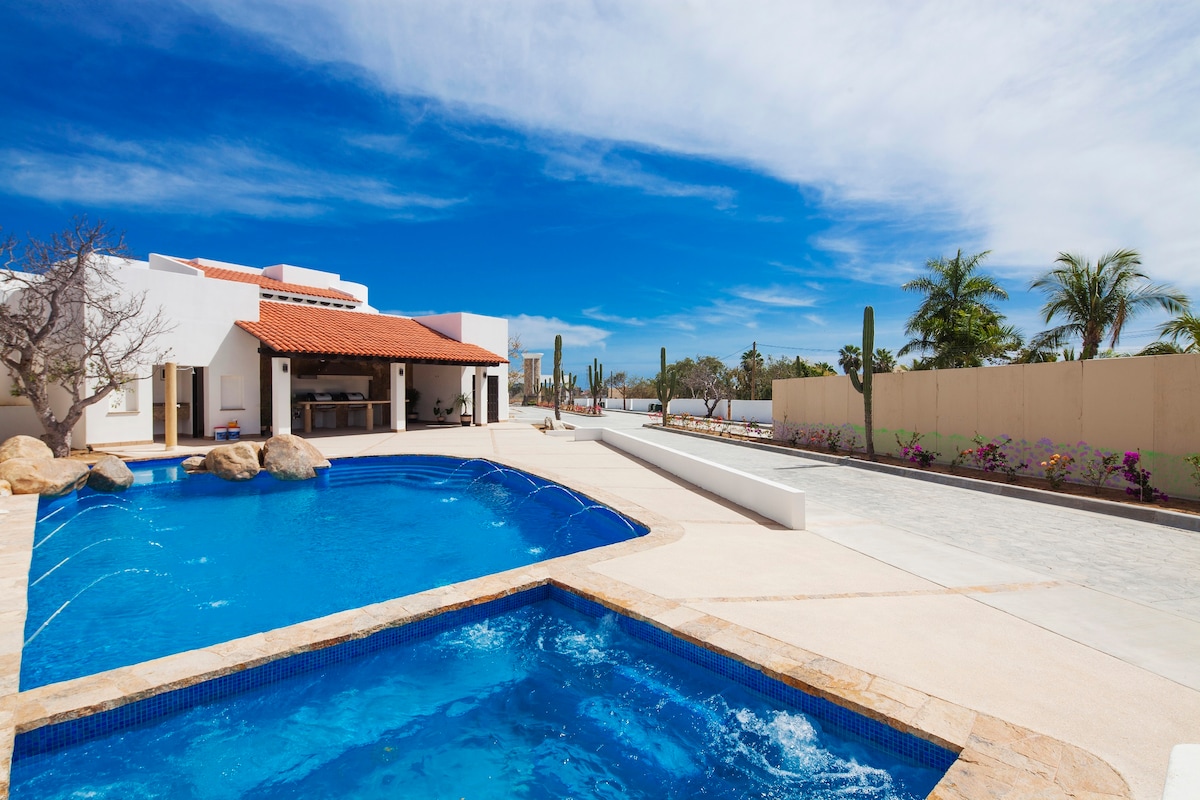 An inviting outdoor area features a large pool and a separate jacuzzi, both surrounded by smooth stone decking. Lush palm trees and cacti are visible in the background, while a covered barbecue area can be seen to one side, providing a perfect space for outdoor gatherings.