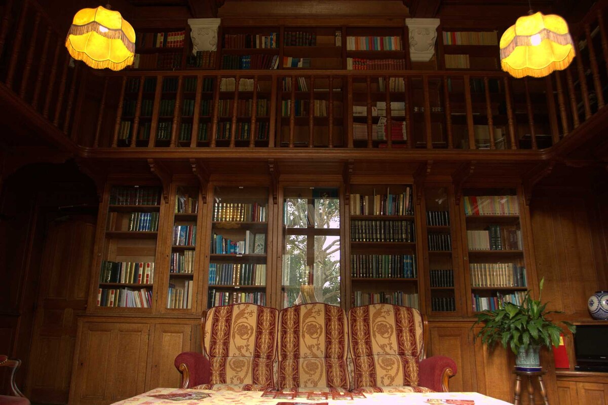 A warm wood-paneled living area features a rich tapestry sofa, complemented by two lamps with a soft glow. The room is lined with bookshelves filled with a variety of books, while a large window allows natural light to illuminate the space, enhancing the inviting atmosphere.