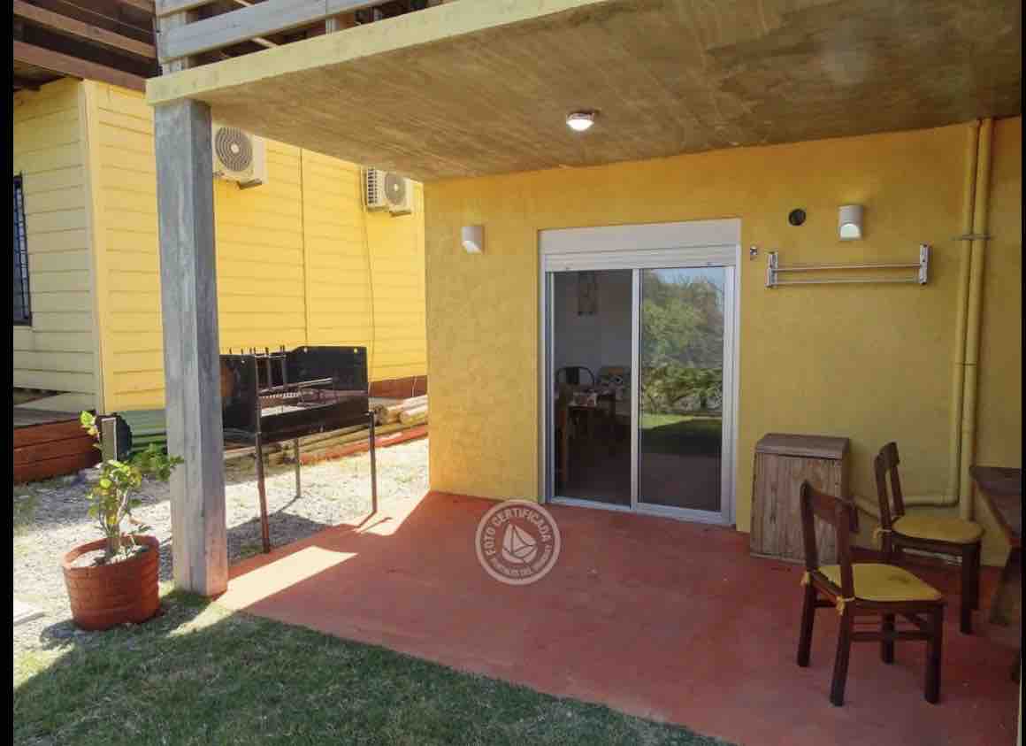 An outdoor area is presented with a covered patio featuring a grill and a small wooden table accompanied by two chairs. A potted plant adds a touch of greenery. The adjacent sliding glass door leads into the interior space.
