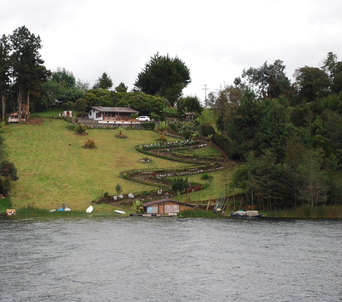 A scenic view of a sloping green hillside is presented, featuring a charming cabin nestled among lush trees and well-maintained flower beds. The calm waters of the lagoon reflect the greenery, with kayaks and canoes visible along the shoreline.