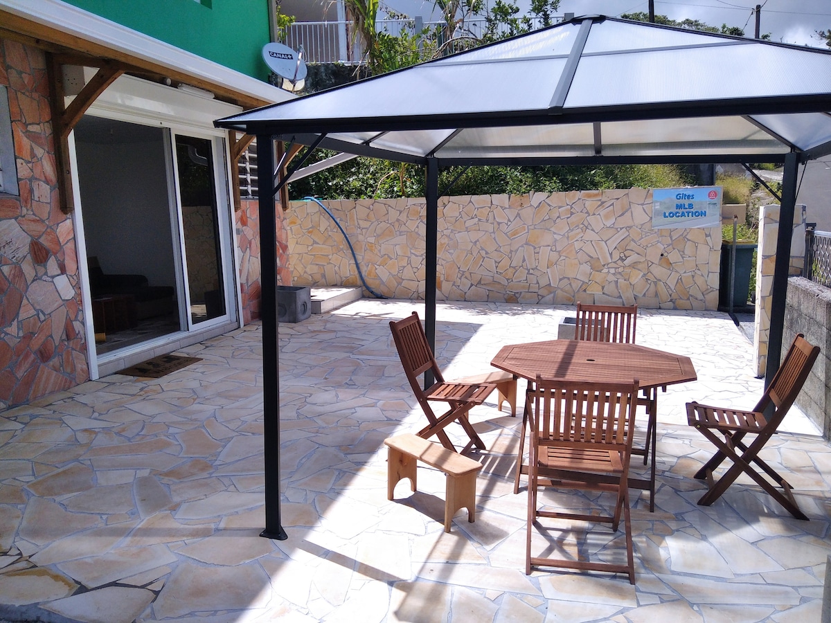 An outdoor patio area features a wooden table surrounded by four folding chairs. A small stool is positioned beside the table. A gazebo provides shelter, while a stone wall serves as a backdrop, contributing to the space's relaxing ambiance.