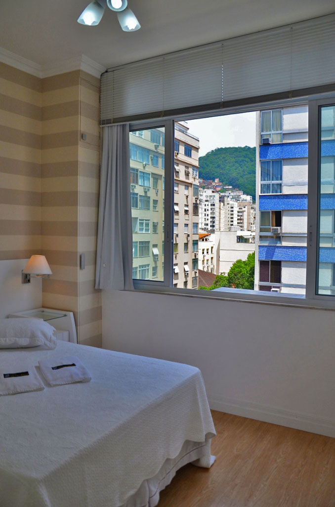 A bright bedroom features a spacious bed dressed in white linens, positioned near a large window. The window offers a view of nearby buildings and a green hillside, allowing natural light to fill the room.