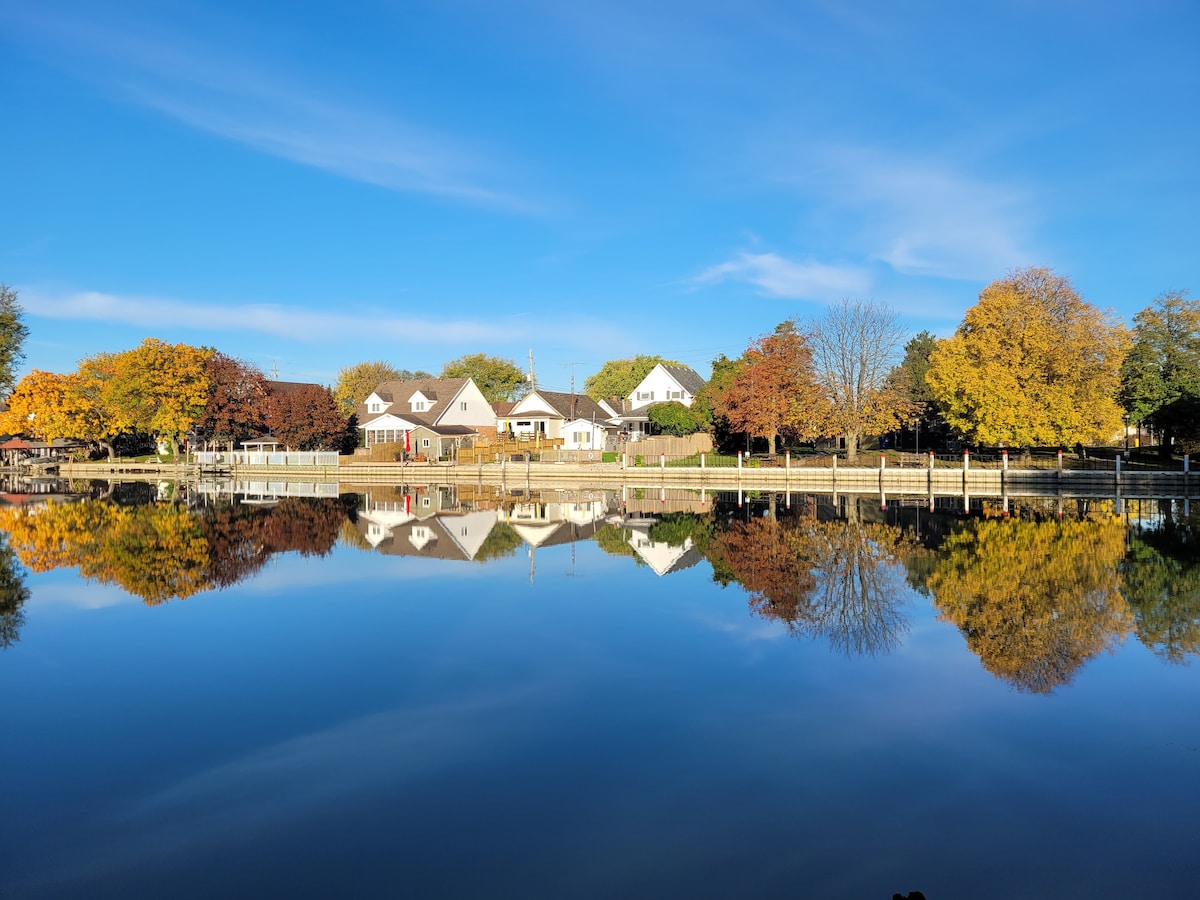 Calm waters reflect charming homes and colorful trees along the riverbank on a clear day. The scene showcases vibrant autumn foliage, with hues of orange, yellow, and green abundant in the trees, creating a serene atmosphere.