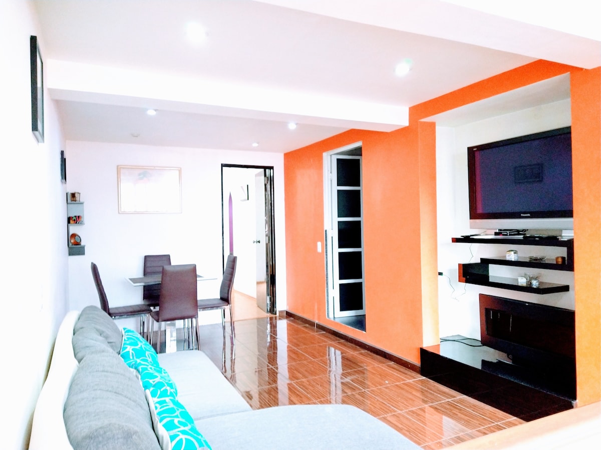 A living area is shown featuring a light-colored sofa with decorative cushions. A small dining table with four chairs is placed near the entrance. An entertainment unit with a flat-screen TV is mounted on an orange accent wall, while lighting fixtures illuminate the space.