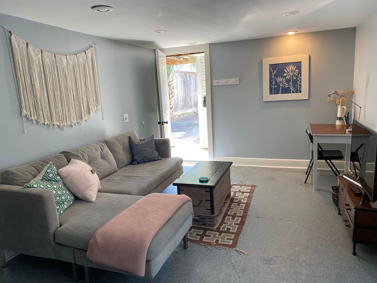 A cozy living area features a gray sectional sofa adorned with a decorative pillow and a soft pink throw. A wooden coffee table sits at its center. Light enters from the door leading to the outside, while a modest desk and framed art enhance the space.