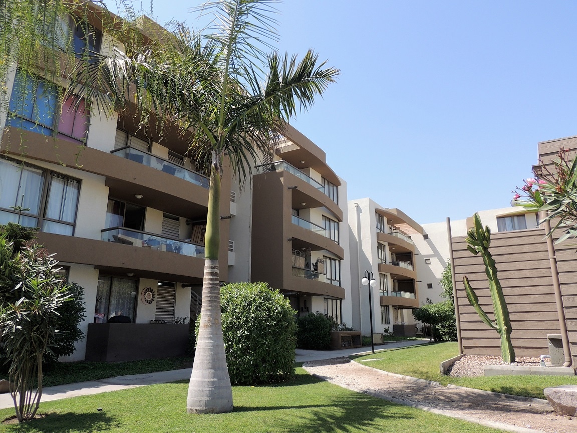 The exterior of a three-story apartment building is displayed, featuring a contemporary design with balconies. Lush greenery and palm trees surround the property, contributing to a peaceful atmosphere. The pathway leads through neatly maintained lawns towards the entrance, inviting exploration.