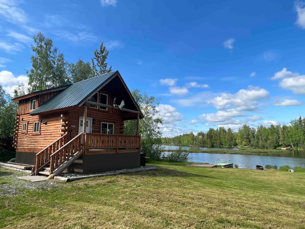 A rustic cabin is positioned next to a serene lake, showcasing a sloped roof and wooden exterior. Lush greenery surrounds the property, and a spacious lawn leads to the water. Soft clouds drift in a blue sky above, enhancing the peaceful lakeside atmosphere.
