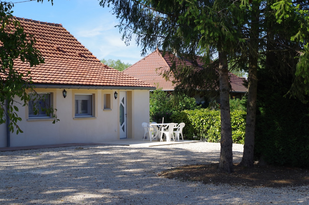 A single-story house with a red-tiled roof is set among greenery. White outdoor furniture is arranged on a gravel patio, surrounded by neatly trimmed hedges and tall trees. The facade features multiple windows that allow natural light into the interior.