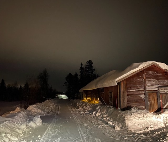 A snow-covered pathway leads to a rustic wooden building adorned with soft lights, creating a welcoming sight in the evening darkness. The surrounding landscape features snow-draped trees, enhancing the serene atmosphere of the peaceful setting.
