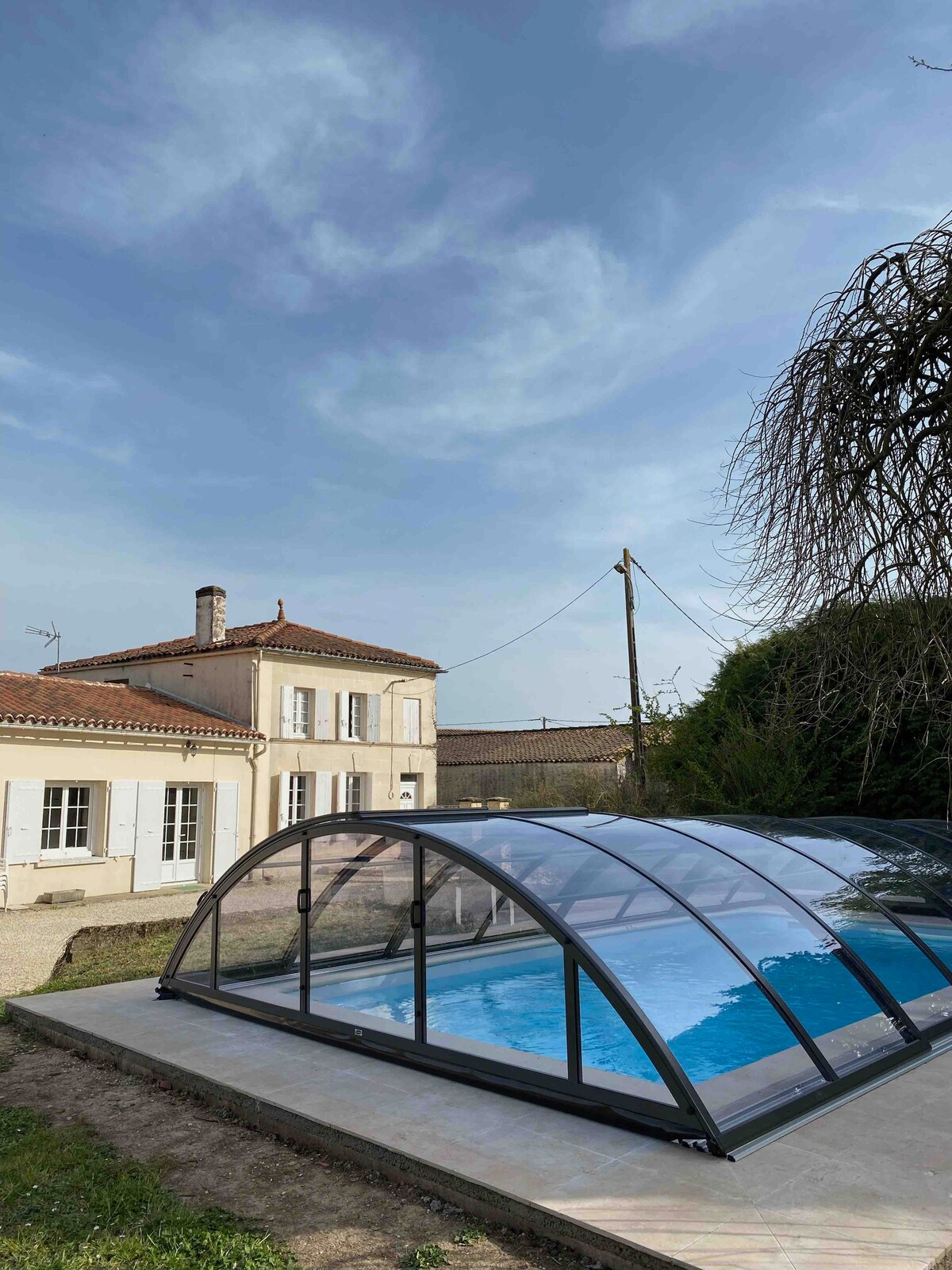 A private pool is enclosed by a transparent cover, providing protection while allowing natural light to illuminate the water. The charming house, featuring traditional architecture, is situated in the background against a clear sky, surrounded by lush greenery.