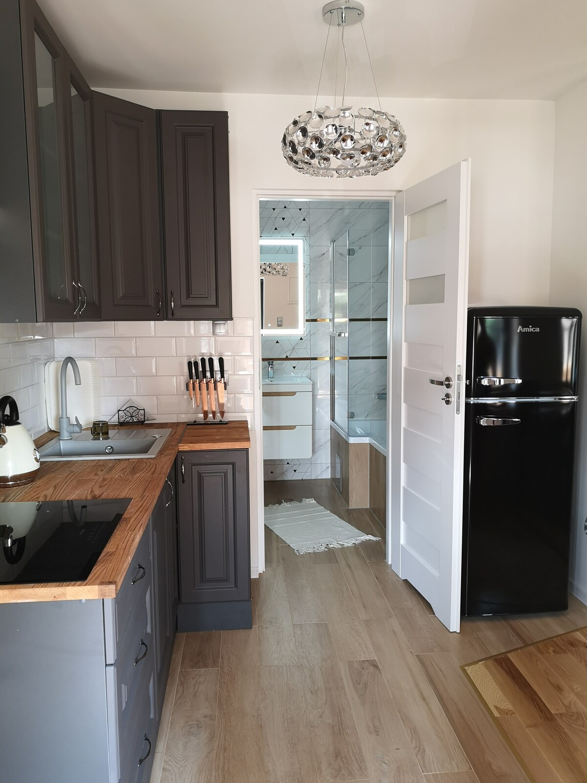 A compact kitchen is equipped with modern appliances and sleek cabinets in dark tones. A counter with wooden accents is featured alongside a refrigerator. Light reflects off a decorative pendant light above, while a doorway leads to a clean bathroom visible in the background.