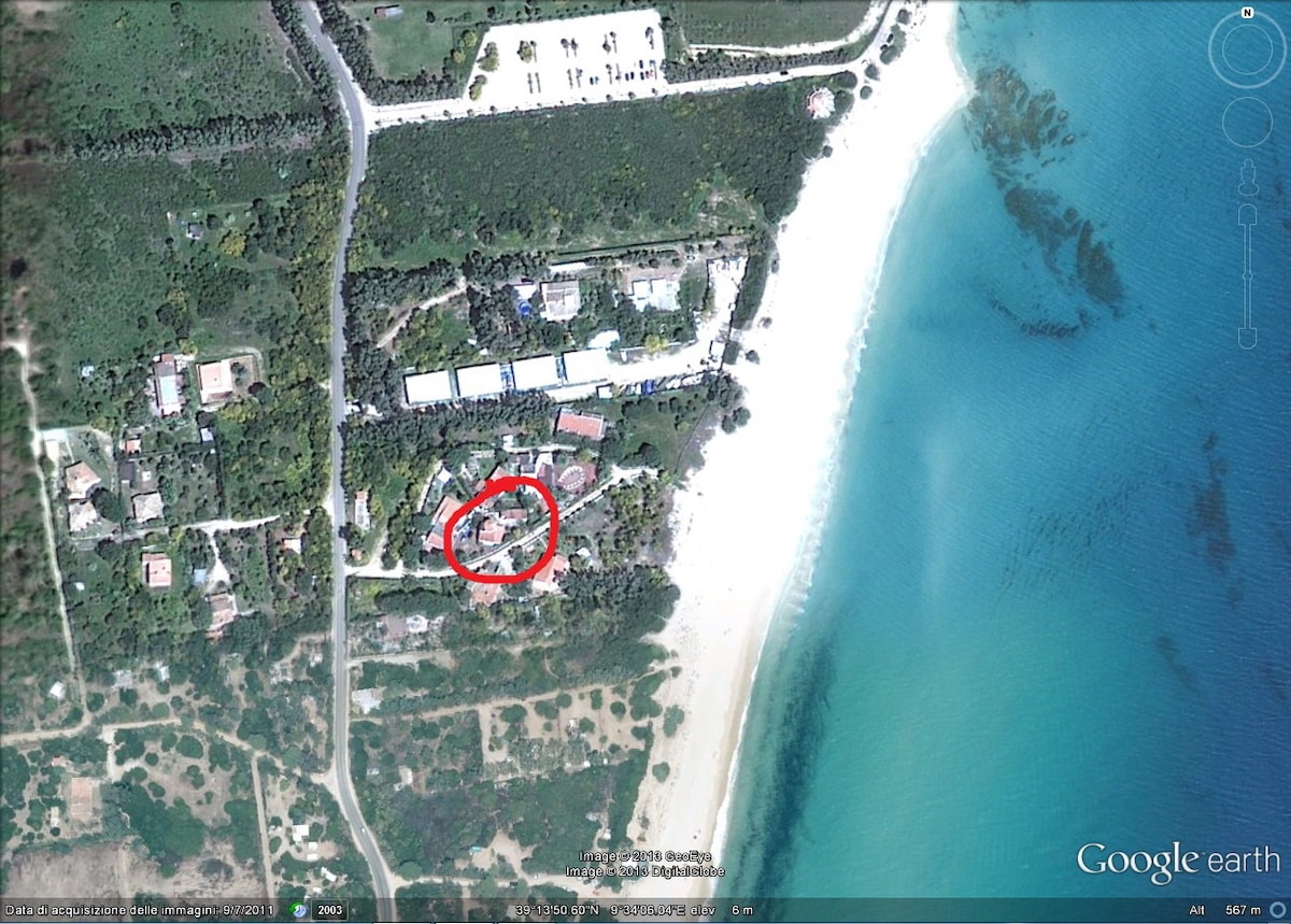 An aerial view highlights the location of the property, surrounded by lush greenery and the coastline. The arrangement of the houses within the small village is visible, along with the nearby sandy beach and clear blue waters.