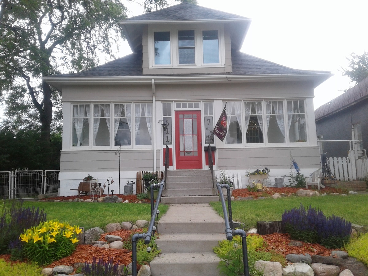 A charming two-story house features a prominent red door and multiple large windows adorned with sheer curtains. Short stone pathways lead through well-maintained flower beds of yellow and purple blooms. The front yard is bordered by a white picket fence, adding to its inviting appearance.
