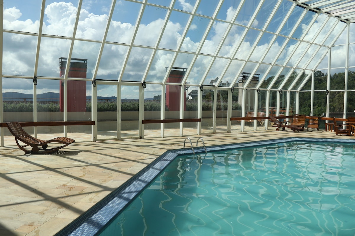 An indoor heated pool is situated within a glass enclosure, allowing natural light to illuminate the space. Lounge chairs are positioned around the pool's edge, with a view of the surrounding mountains visible through the transparent walls.