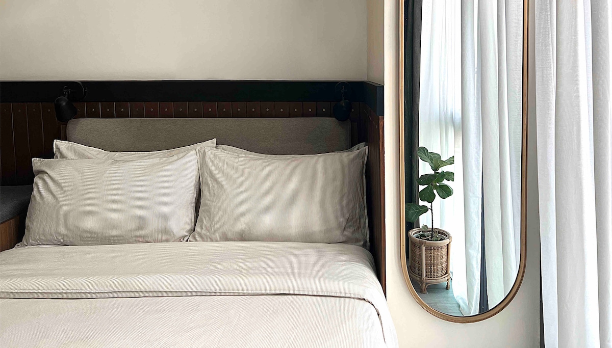 A spacious bed is dressed in soft, neutral-toned linens, complemented by a pair of plush pillows. A decorative mirror hangs next to a window draped with light curtains, revealing a potted plant placed on the windowsill.