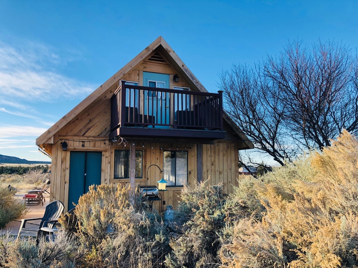 The exterior of the Desert Sage Chalet is framed by the natural beauty of desert sagebrush. The charming wooden structure features a sloped roof and a balcony, where guests can enjoy scenic views. The front door is painted in a contrasting color, welcoming visitors.