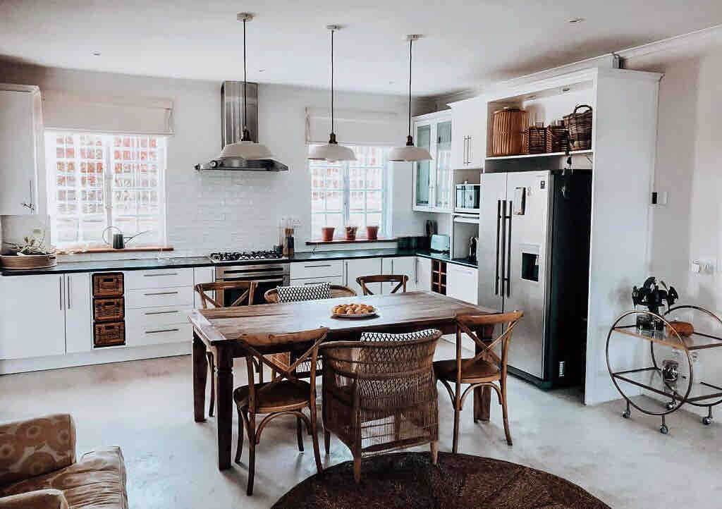 A spacious kitchen area features a large wooden dining table surrounded by wicker chairs. Modern appliances, including a black refrigerator and a gas stove, are integrated into the bright cabinetry. Natural light fills the room through two windows, enhancing the open and functional design.