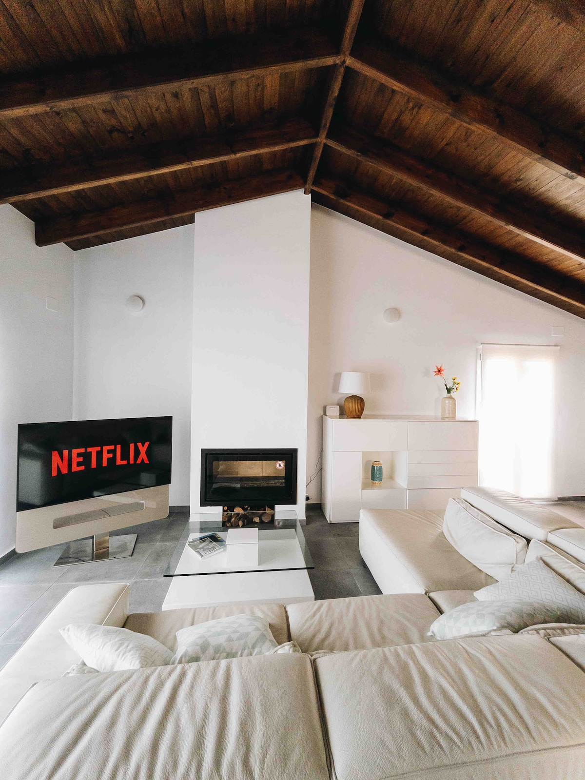 A spacious living area features a modern design with high wooden ceilings. A large sectional sofa is positioned in front of a minimalist fireplace. A flat-screen TV displays a streaming service logo. Natural light filters through the nearby window, enhancing the inviting atmosphere.