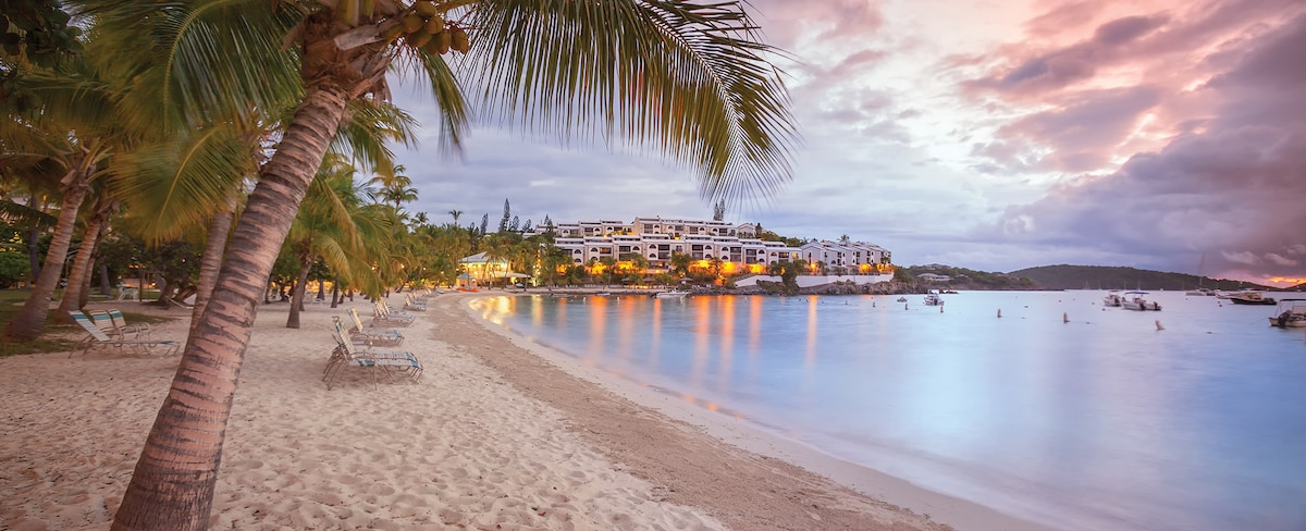 The image showcases a serene beachfront at sunset, with soft sand gently meeting the calm water. Palm trees frame the scene, while a multi-level resort sits in the background, illuminated by warm lighting. Small boats are visible on the water's surface.
