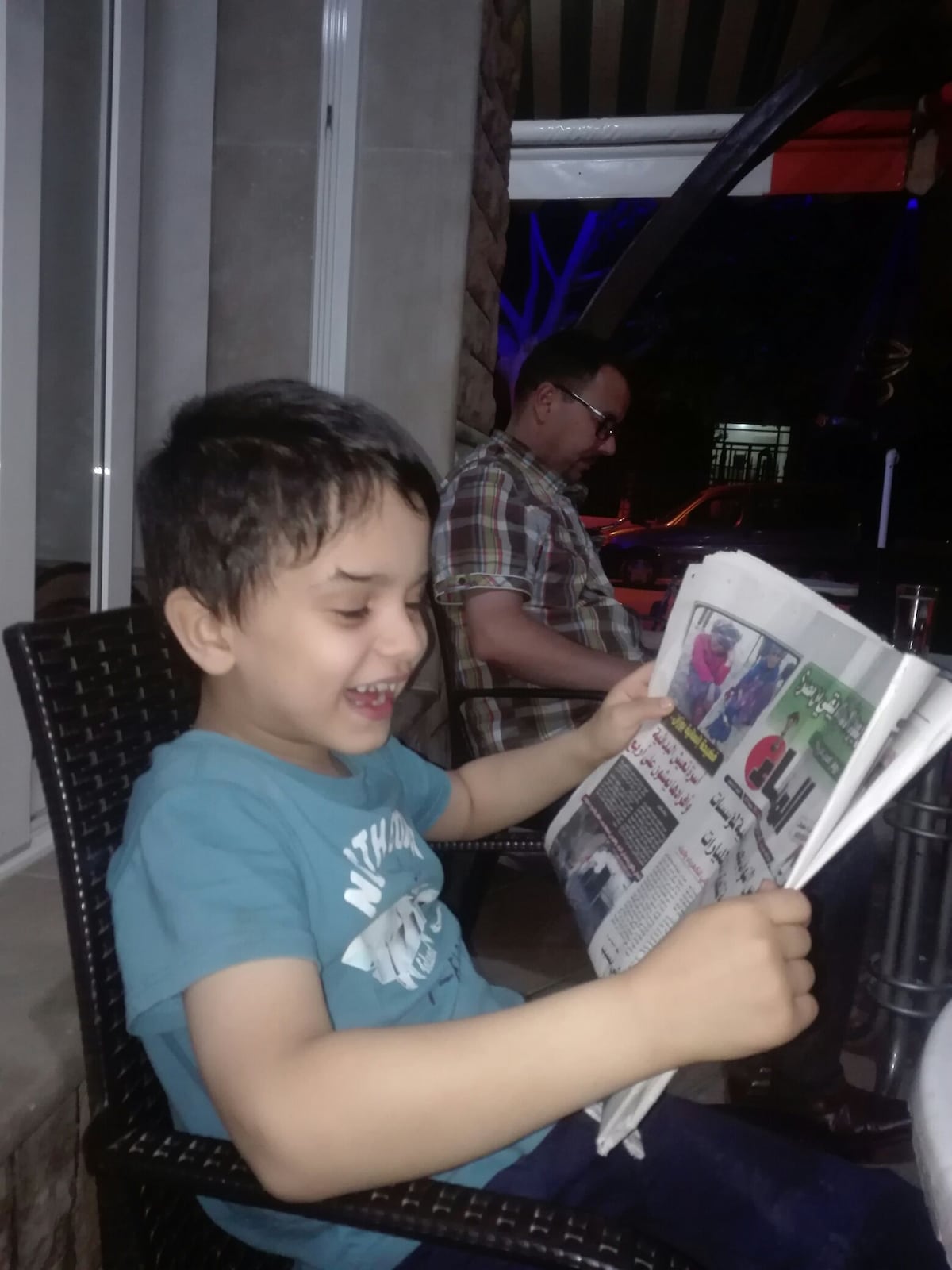 A young child in a light blue shirt is seated at an outdoor table, holding a newspaper. The setting includes a stone wall and striped awning, with dim lighting providing a relaxed atmosphere. A second figure can be seen in the background, seated and engaged in a casual conversation.