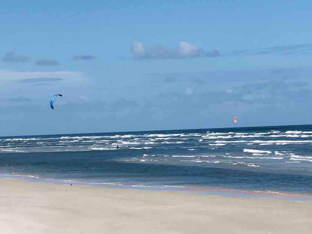 The image captures a serene coastal scene featuring a sandy beach and expansive ocean view. Gentle waves create a rhythmic pattern against the shore, while colorful kites can be seen soaring in the bright blue sky, adding a sense of activity to the tranquil setting.