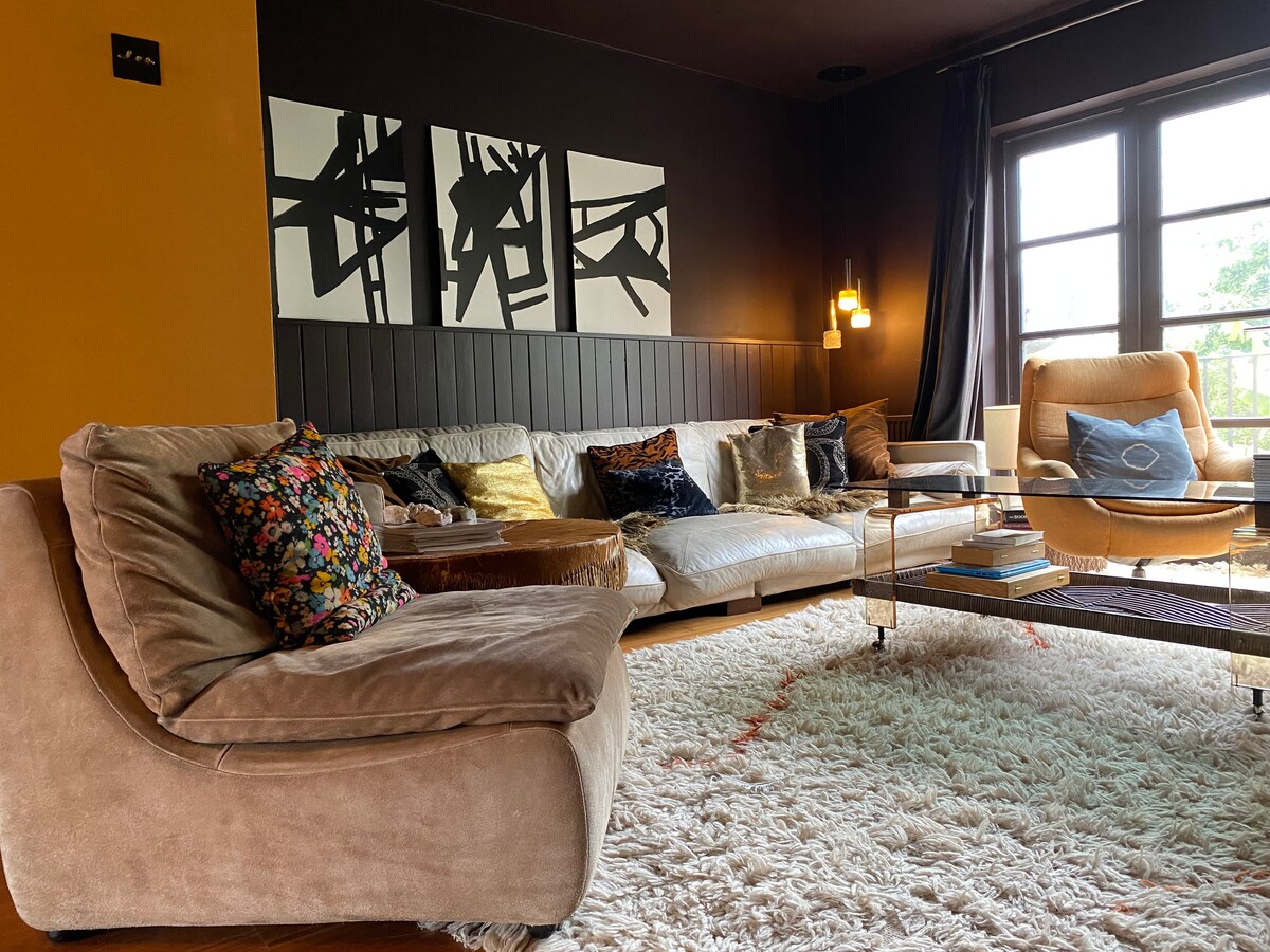A cozy sitting room is furnished with a large, curved sofa adorned with decorative cushions. A glass coffee table sits atop a plush, white area rug. The space is illuminated by stylish pendant lighting, and large windows offer views of the outside.