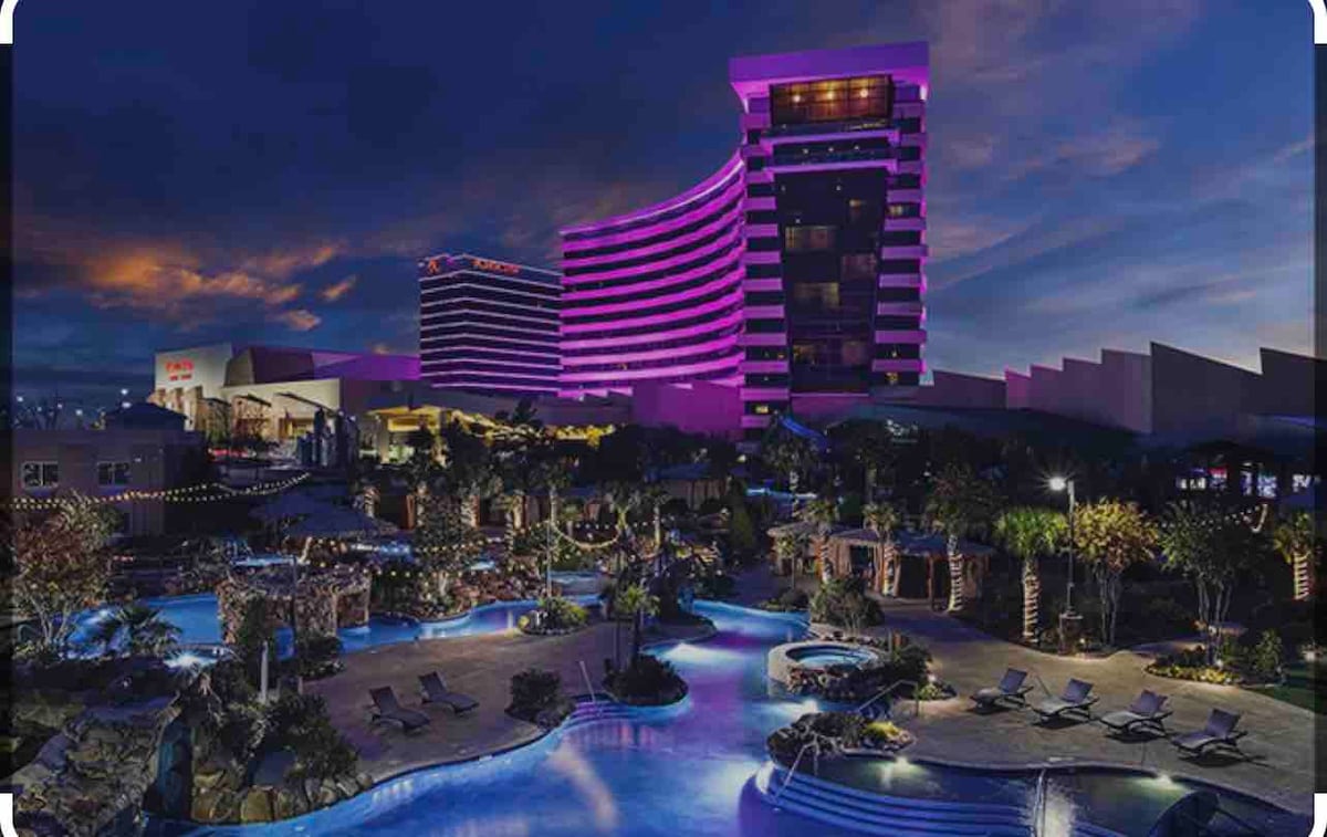 A large, modern hotel overlooks a vibrant outdoor area with pools and lounge chairs. The structure is illuminated in purple lighting against a darkening sky, while landscaped palm trees and various seating options create a welcoming atmosphere.