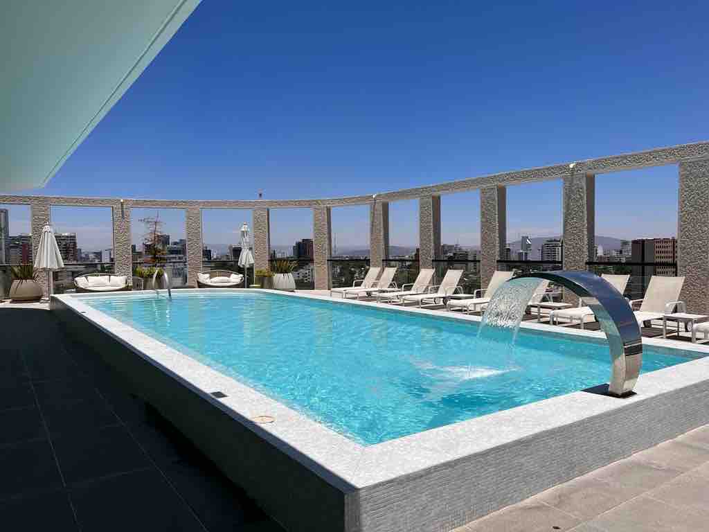 An outdoor pool area is shown, featuring clear blue water and a sleek design. Sun loungers are arranged around the pool, and modern architectural details frame the space. A water feature cascades elegantly into the pool, while panoramic views of the city are visible in the backdrop.
