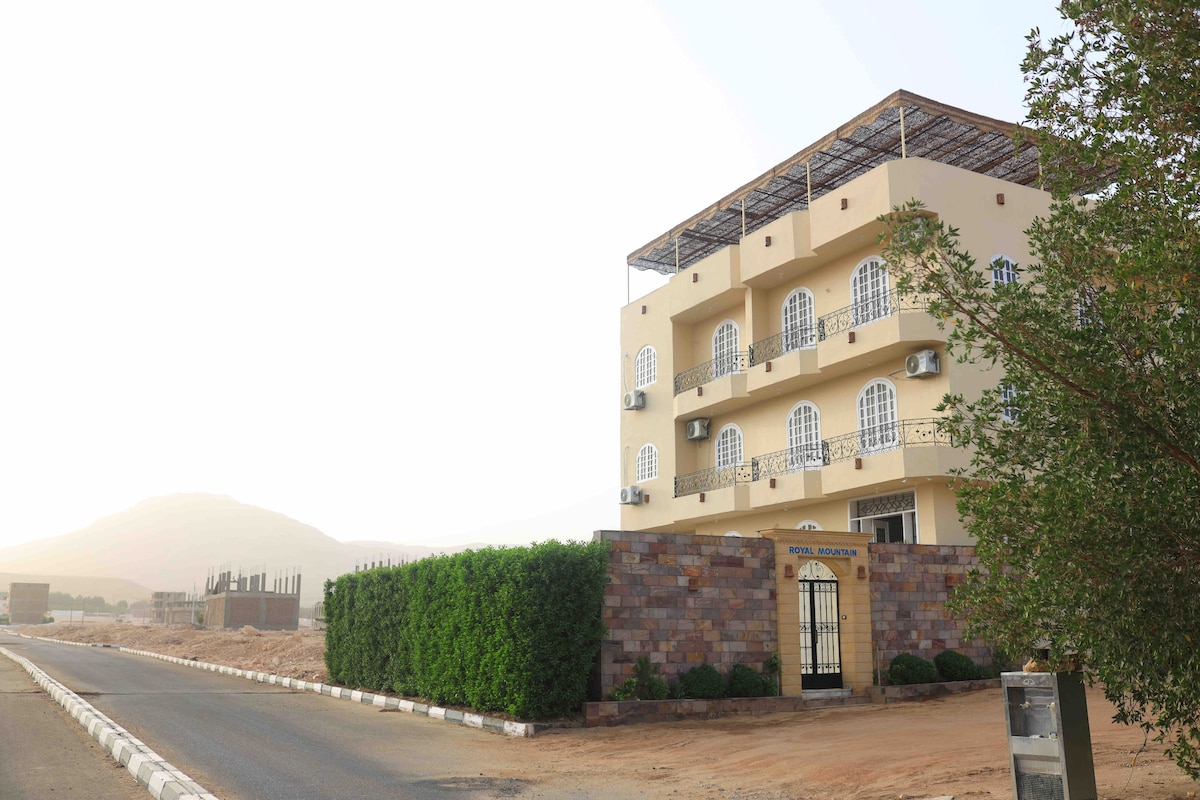 The Royal Mountain Hotel is shown from the street, featuring a three-story structure with arched windows and a rooftop terrace. A stone wall encloses the property, with neatly trimmed greenery and a pathway leading to the main entrance. Surrounding areas are visible, including distant hills.