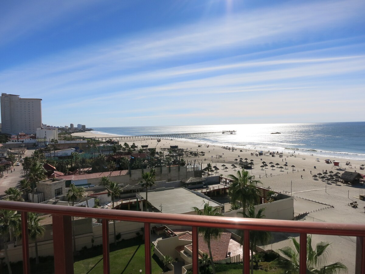 A panoramic view of the ocean and beach is captured from the top floor. Sunlight reflects off the water, creating a shimmering effect. The sandy beach is dotted with umbrellas and lounge chairs, while palm trees frame the lower part of the image, complementing the coastal scenery.