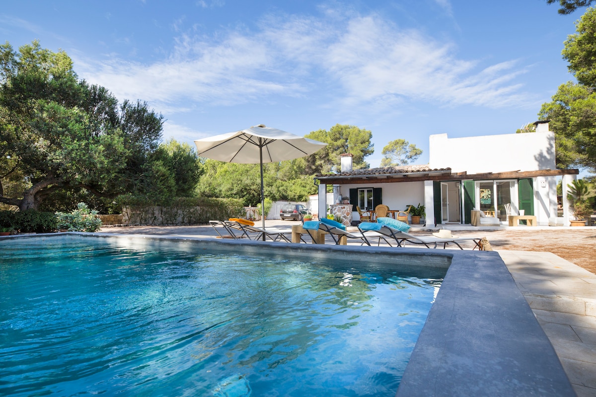 The image features a private pool surrounded by sun loungers and an umbrella, set against a backdrop of lush pine trees. The house, with large glass doors, has a simple, modern design. A spacious terrace extends from the house, inviting relaxation outdoors.