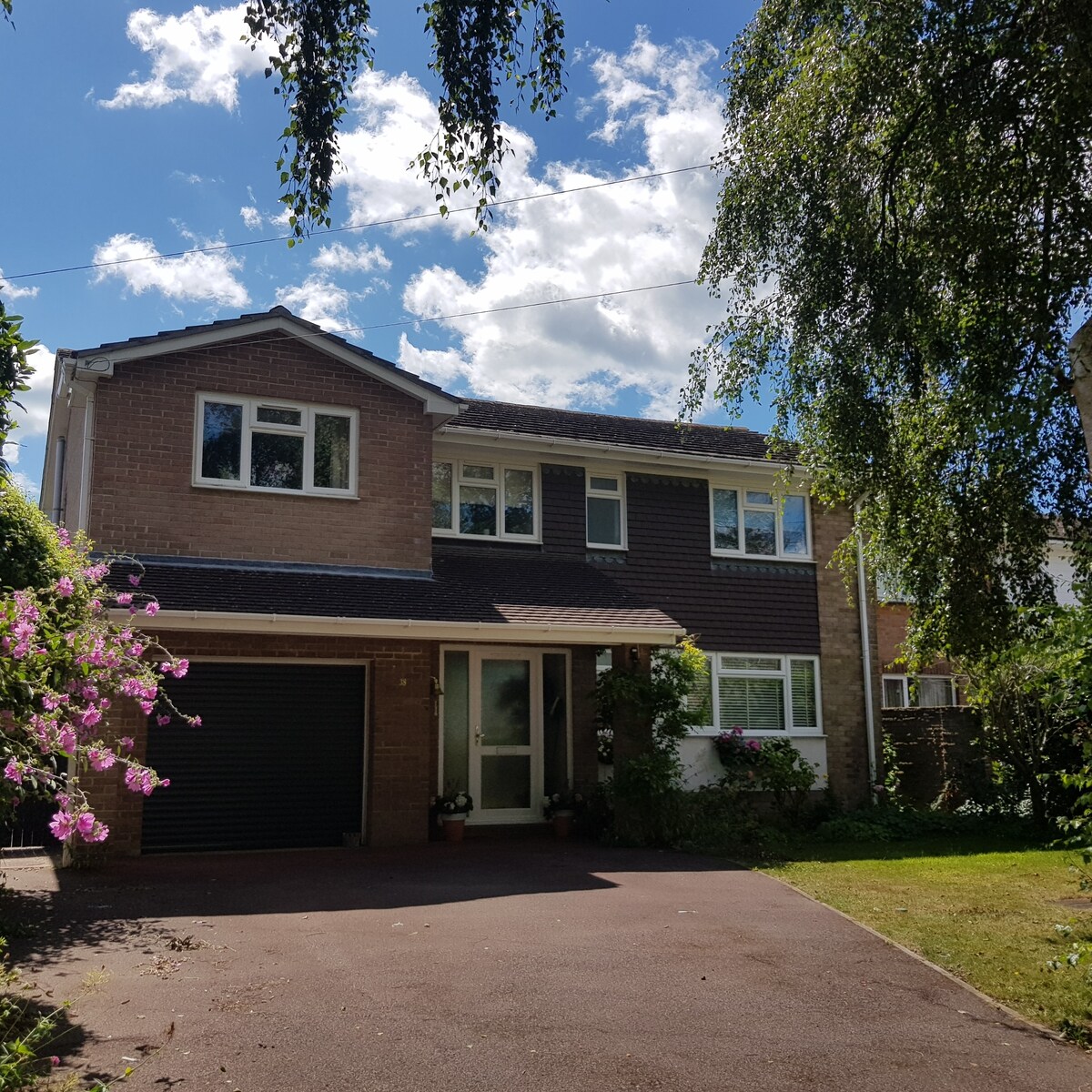 A two-story house is shown, featuring a mix of brick and wood siding. The front entrance includes a welcoming doorway surrounded by greenery and flowering plants. A paved driveway leads to a garage, and the sky is partly cloudy, illuminating the exterior.