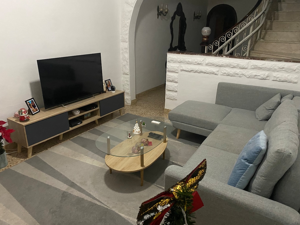 A modern living area is highlighted by a large television positioned on a low wooden cabinet. A sectional couch in soft gray contrasts with a light area rug. A round coffee table sits in front, while decorative items add a personal touch to the space.
