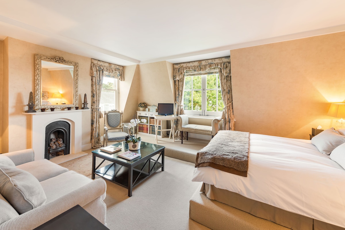 A spacious room features neutral-toned walls and soft carpet. A comfortable seating area is arranged with a plush sofa, coffee table, and decorative plant. Natural light filters through large windows, illuminating the cozy bedding and tasteful decor. A traditional fireplace adds character to the space.