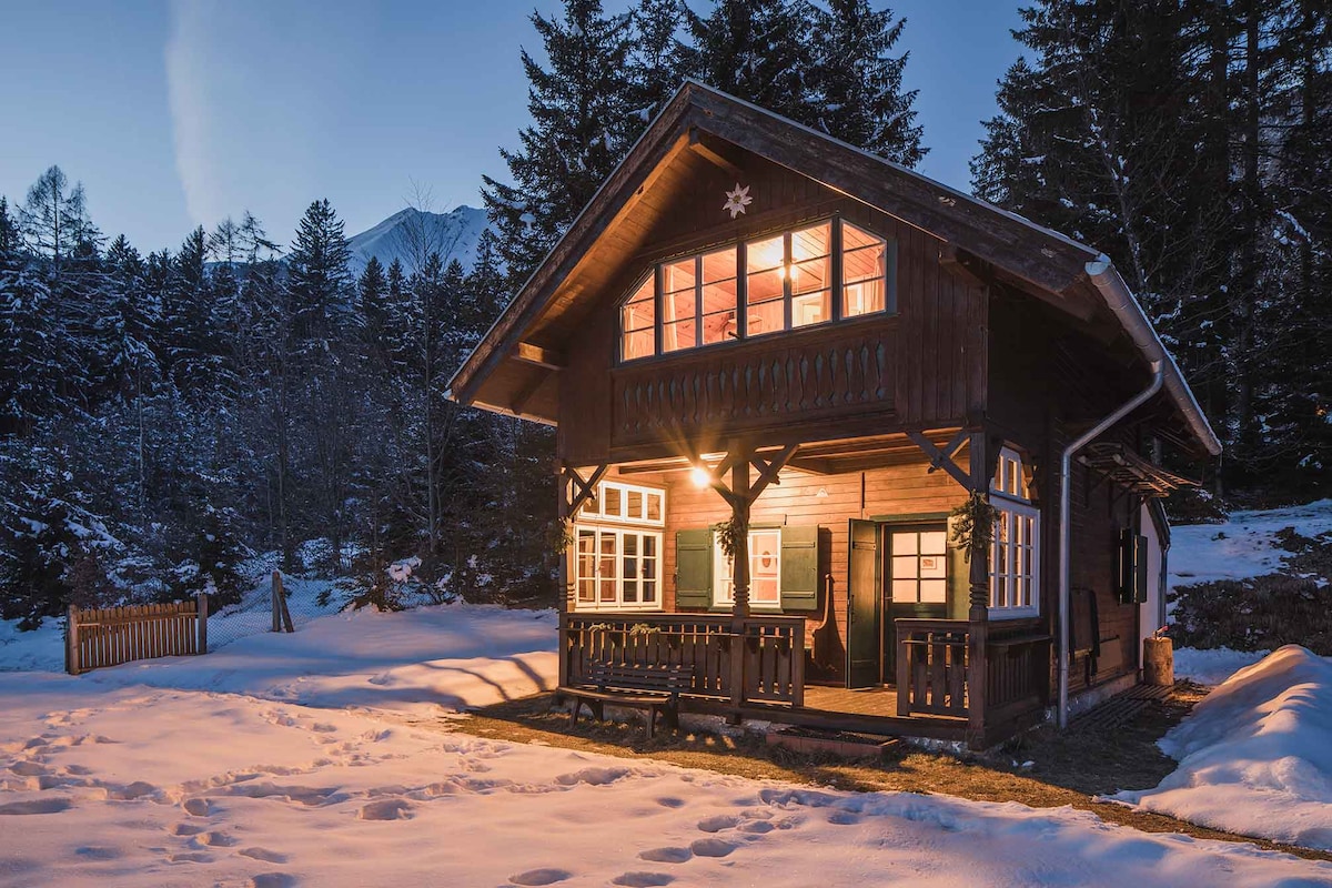 A historic chalet is set against a snowy landscape, framed by tall trees. Warm light glows from the windows, illuminating the wooden exterior. The front porch features a bench, inviting relaxation in the serene surroundings. Snow blankets the ground, enhancing the cabin's cozy appearance.
