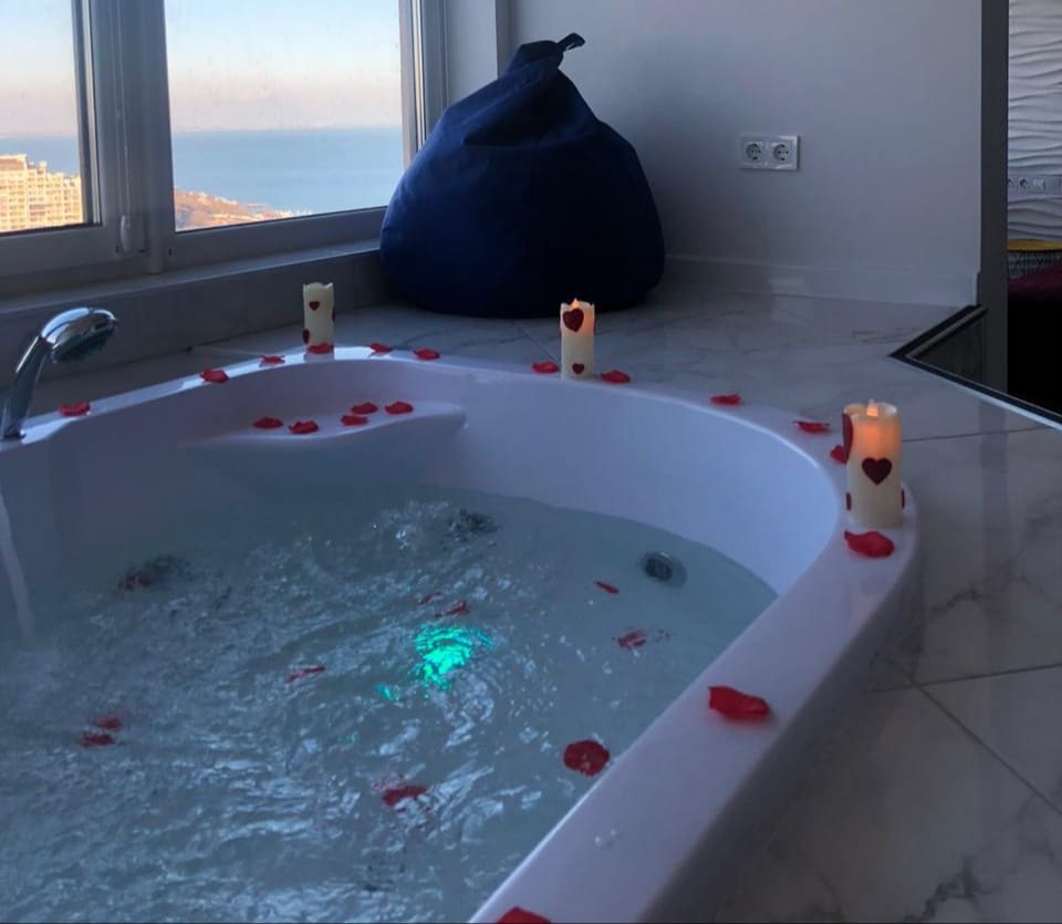 A modern jacuzzi is positioned near a window, offering a view of the sea. The tub is adorned with rose petals and surrounded by candles, creating a serene ambiance. Natural light reflects off the water, enhancing the relaxing atmosphere.
