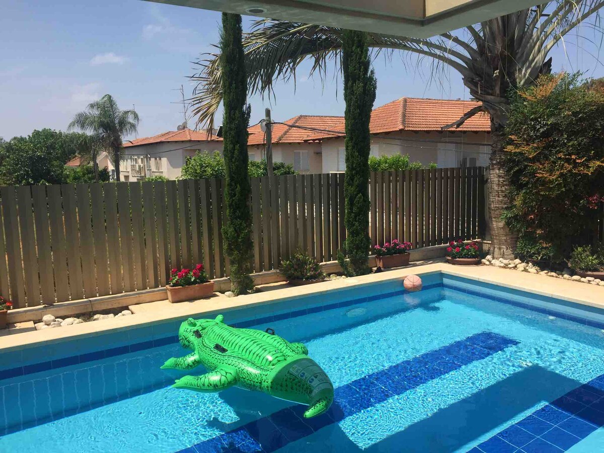 A swimming pool is showcased with a green inflatable crocodile resting on the surface. Peaceful views of the surrounding garden are visible, featuring palm trees and potted flowers along a fence that provides added privacy.