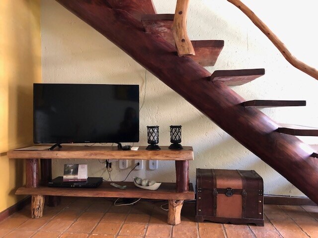 A wooden staircase with a natural finish is featured alongside a rustic TV stand made of reclaimed wood. A flat-screen television is positioned on the stand, with decorative lanterns and a vintage trunk placed nearby, contributing to a cozy ambiance.