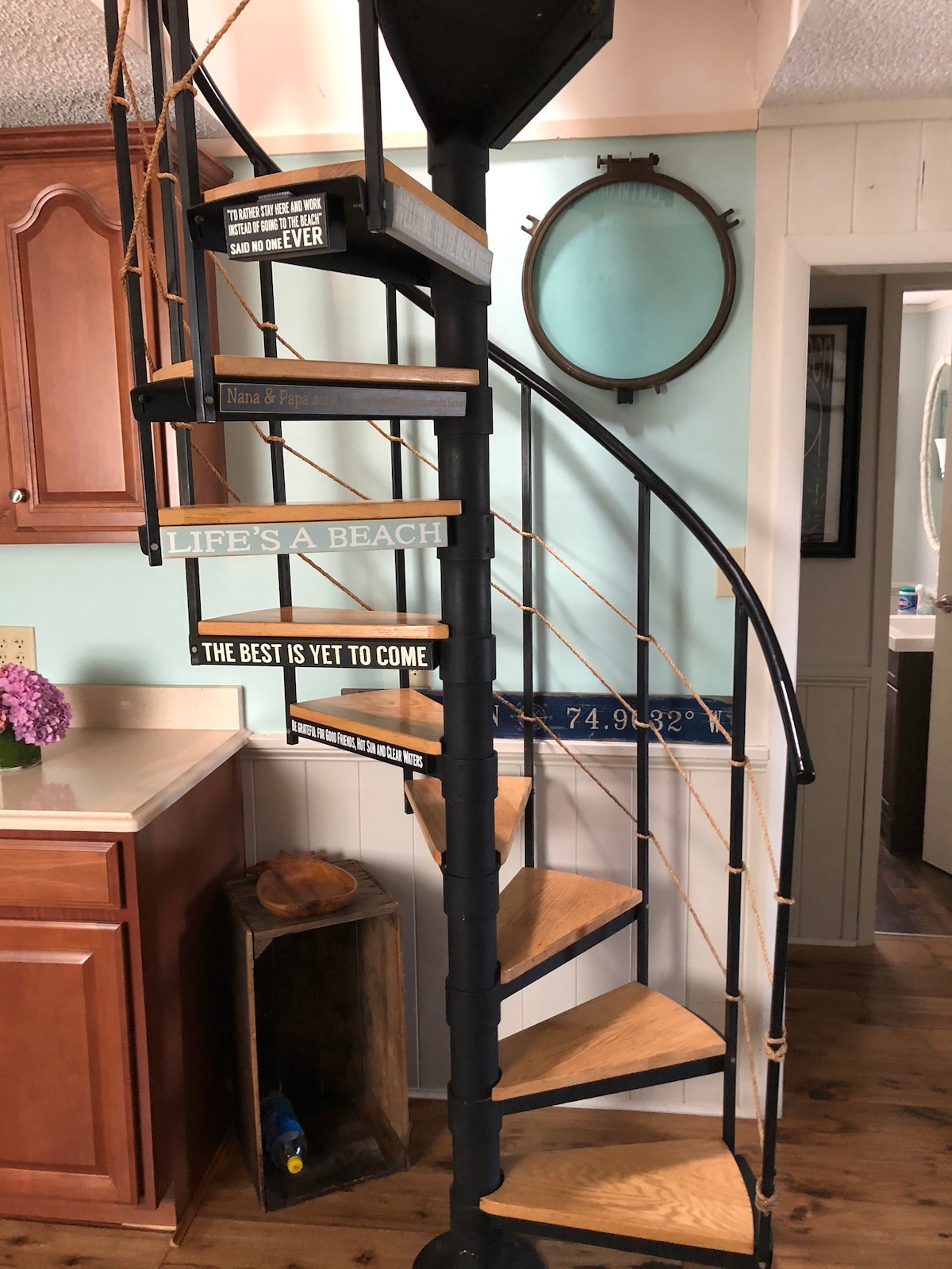 A spiral staircase with wooden steps and metal railings is featured, leading to additional living spaces. The wall nearby is adorned with a round mirror and a small bouquet of flowers. Inspirational signs decorate the staircase, enhancing its playful charm.