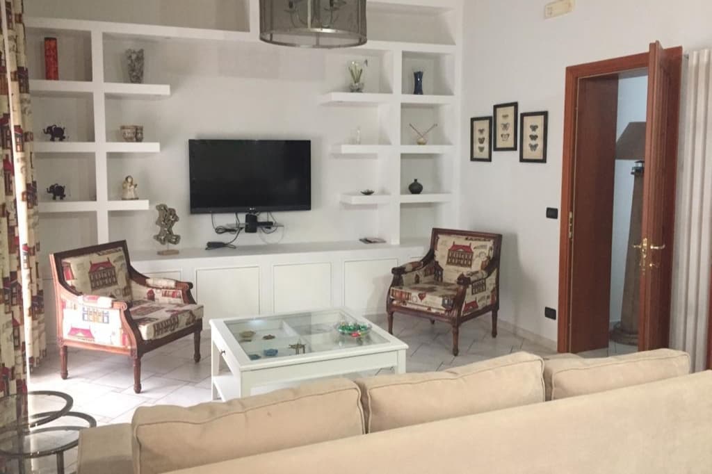 An inviting living room features a comfortable beige sofa and two patterned armchairs, arranged around a glass coffee table. A wall-mounted television is set against a white shelving unit, which displays decorative items. Natural light enters through the windows, enhancing the spacious ambiance.